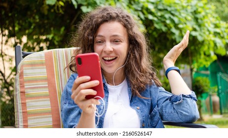 Excited curly girl looking at mobile phone surprised by good unbelievable news, feeling amazed, sitting in green garden on backyard.Good news,great result, lottery win,new opportunity, get new job