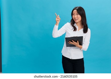Excited cute Asian businesswoman in classic office dress code holds folder tablet with resume point finger up posing isolated on over blue studio background. Cool business offer. Job interview concept
