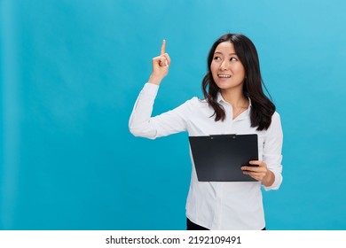 Excited Cute Asian Businesswoman In Classic Office Dress Code Holds Folder Tablet With Resume Point Finger Up Posing Isolated On Over Blue Studio Background. Cool Business Offer. Job Interview