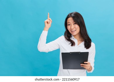 Excited Cute Asian Businesswoman In Classic Office Dress Code Holds Folder Tablet With Resume Point Finger Up Posing Isolated On Over Blue Studio Background. Cool Business Offer. Job Interview Concept