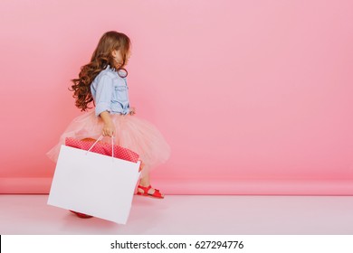 Excited cute little girl in tulle pink skirt, with long brunette hair walking with big white package isolated on pink background. Enjoying shopping in childhood, expressing positivity. Place for text