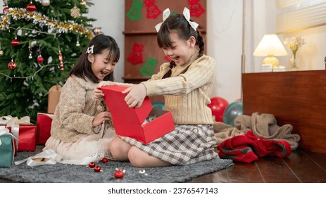 An excited, cute young Asian girl is opening her Christmas gift with her younger sister near the Christmas tree in the living room, having fun and celebrating Christmas at home together.