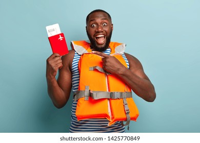 Excited Dark Skinned Guy Points At Passport With Tickets, Has Unexpected Trip Abroad, Wears Safety Lifejacket, Has Muscular Arms, Poses Over Blue Background. People, Journey, Vacation Concept