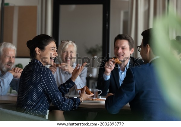Excited diverse employees eating pizza during break\
in office together, happy Indian businesswoman laughing at funny\
joke, talking chatting with colleagues, having fun, sharing\
corporate lunch