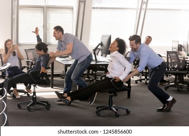 Excited Diverse Employees Having Fun Together In Office, Riding On Chairs At Work, Enjoying Break, Laughing, Colleagues Shoot Video On Phone, Engaged Funny Activity, Celebrating Corporate Success