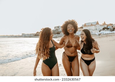 Excited diverse female friends in swimwear embracing and laughing cheerfully while walking at the beach, enjoying their free time on ocean shore
