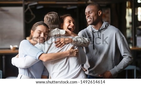 Excited diverse friends embrace greeting male buddy coming at meeting in cafe, multiracial students group hugging guy laughing say hello welcoming at students reunion, multi ethnic friendship concept