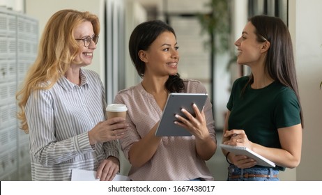 Excited diverse multiethnic female colleagues stand brainstorming discussing ideas using modern tablet gadget, happy multiracial woman coworkers employees cooperate working on pad in office