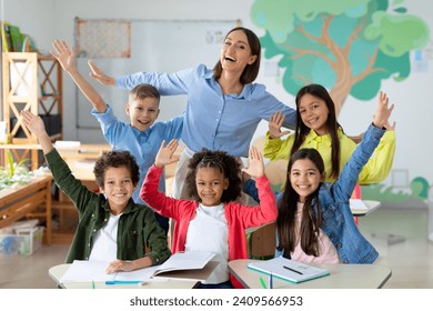 Excited diverse schoolchildren raising arms and smiling, sitting in classroom with their happy female teacher - Powered by Shutterstock