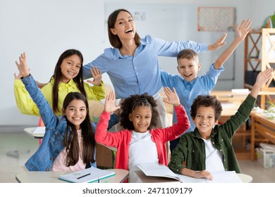 Excited diverse schoolchildren raising arms and smiling, sitting in classroom with their happy young female teacher, back to school concept - Powered by Shutterstock