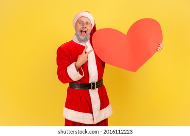 Excited elderly man with gray beard wearing santa claus costume keeps mouth open, showing big red heart and v sign, celebrating holiday. Indoor studio shot isolated on yellow background.