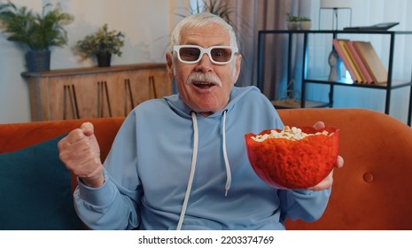 Excited Elderly Man On Sofa Eating Popcorn And Watching Interesting Tv Serial, Sport Game, Film, Online Social Media Movie Content Online At Home. Senior Grandfather Enjoying Domestic Entertainment