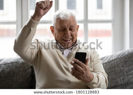 Excited elderly mature retired man looking at telephone screen, celebrating online lottery win or getting message with good news. Emotional mature 70s grandfather showing yes gesture, laughing indoors