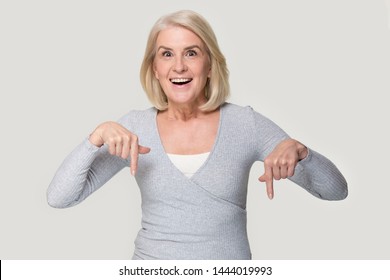 Excited Elderly Woman Head Shot Showing With Her Fingers Down. Happy Smiling Old Female In Years Point By Finger To Bottom Looking At Camera. Aged Person Studio Portrait Isolated On Gray Background