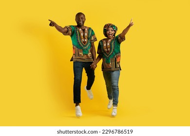 Excited emotional african couple wearing traditional costumes holding hands, running and pointing at camera, chasing great deal, yellow studio background, full length, copy space - Powered by Shutterstock