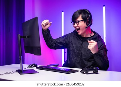 Excited Face Of Asian Gamer With Headphone Playing Computer Pc Video Game Online Sitting At Living Room. Young Professional Gamer Streaming On Social Playing Game Very Fun