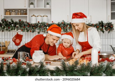 Excited Family Mother, Father And Son In Santa Hats Cutting Dough With Cookie Cutters On A Messy Kitchen Table Over Christmas Lights