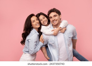 Excited family of three having fun on pink background, girl piggybacking dad, happy young parents and their daughter smiling at camera - Powered by Shutterstock