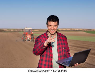 Excited Farmer Holding Laptop And Pointing Finger At Camera In Front Of Tractor In Field