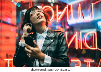 Excited fashion female lover of music dressed in stylish leather jacket listening songs online in earphones connected to smartphone while enjoying night lights and neon illumination in New York City - Powered by Shutterstock