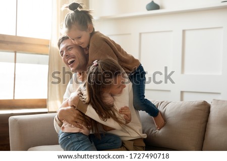 Excited father having fun with two little daughters, spending weekend at home, sitting on comfortable couch, laughing cute preschool girls and dad hugging, cuddling, enjoying free time together