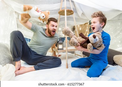 Excited Father And Son Sitting In Blanket Fort And Playing With Teddy Bears  