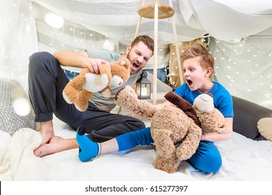 Excited Father And Son Sitting In Blanket Fort And Playing With Teddy Bears  