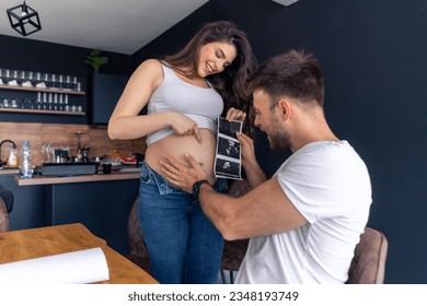 An excited father-to-be holds an ultrasound to his partner's stomach and smiles because he is looking forward to having a baby soon - Powered by Shutterstock