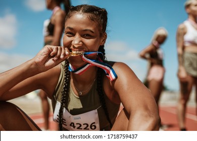 Excited female athlete biting her medal while sitting on race track with other athletes in background. Sports woman enjoying winning a medal in sprinting event at stadium. - Shutterstock ID 1718759437