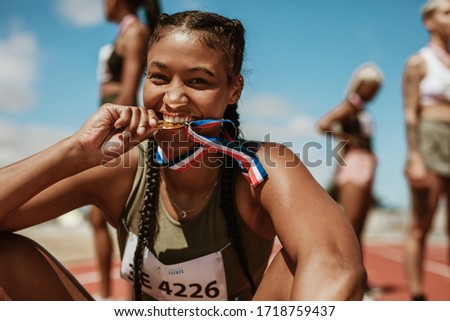 Excited female athlete biting her medal while sitting on race track with other athletes in background. Sports woman enjoying winning a medal in sprinting event at stadium.
