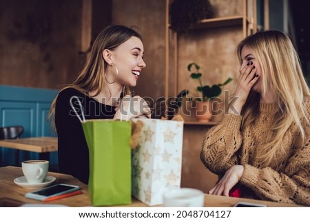 Excited female friends in casual clothes smiling and giving presents to each other while sitting at wooden table in cozy cafe