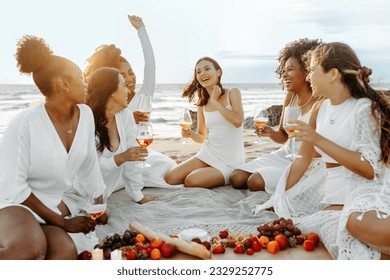 Excited female friends making toast celebration hen party, drinking wine, eating fresh fruits and chatting, having picnic by seashore at sunset - Powered by Shutterstock