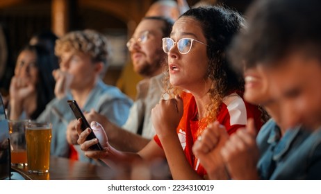 Excited Female Holding a Smartphone, Sitting at a Bar Stand. Nervous About the Sports Bet She Put on a Her Favorite Soccer Team. Joyful When Football Team Scores a Goal and She Wins the Prize. - Powered by Shutterstock