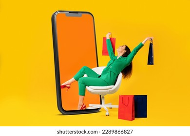 Excited female shopaholic sitting on chair with shopper bags near giant cellphone display over yellow studio background. Cheerful woman enjoying seasonal sales. Collage