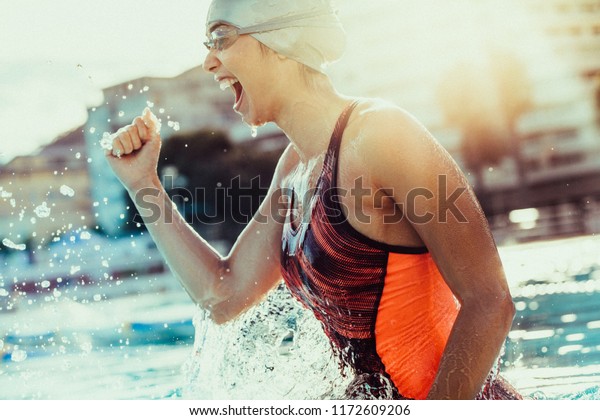 Excited female swimmer with clenched\
fist celebrating victory in the swimming pool. Woman swimmer\
cheering success in pool wearing swim goggles and\
cap.