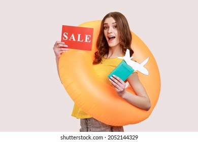 Excited Female Of Young Age With Brown Hair Standing With Orange Rubber Ring, Holding Passport Document And Airplane Mockup, Low Price For Vacation. Indoor Studio Shot Isolated On Gray Background.