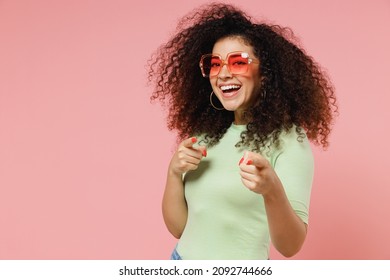 Excited fun young curly latin woman 20s years old wears mint t-shirt sunglasses pointing index fingers camera on you motivating encourage isolated on plain pastel light pink background studio portrait
