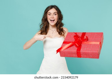 Excited funny bride young woman 20s in white wedding dress pointing index finger on red present box with gift ribbon bow isolated on blue turquoise wall background. Ceremony celebration party concept