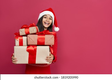 Excited Funny Indian Latin Kid Child Girl Wears Santa Hat Looking At Camera Holding Many Gifts Boxes Celebrating Happy 2022 New Year Isolated On Red Background. Merry Christmas Presents Shopping Sale.