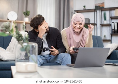 Excited Funny Multiehnic Muslim Couple Having Fun Together While Playing Video Games At Home. Laughing Woman In Hijab Celebrating Victory, While Her Friend Is Sad Because Of Lost.