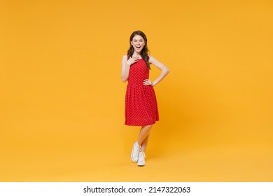Excited funny young brunette woman girl in red summer dress posing isolated on yellow wall background studio portrait. People sincere emotions lifestyle concept. Mock up copy space. Put hand on chest