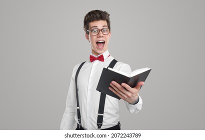 Excited funny young male poet in white shirt with bow tie and suspenders and eyeglasses reading poem written in notebook against gray background
