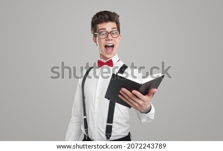 Excited funny young male poet in white shirt with bow tie and suspenders and eyeglasses reading poem written in notebook against gray background