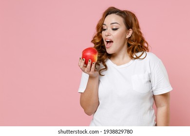 Excited funny young redhead plus size body positive woman 20s wearing white casual t-shirt hold eating ripe fresh apple fruit looking camera isolated on pastel pink color background studio portrait