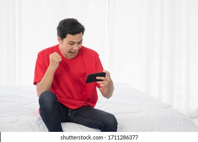 Excited Gamer Male, Happy Young Attractive Asian Man Win Plays Game Online On Smartphone Or Mobile Phone And Triumphant With Raised Fists While Sitting On Bed In The Bedroom At Home.