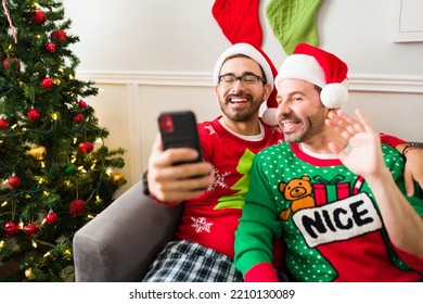 Excited Gay Young Men Saying Hi While Having A Video Call On The Smartphone With Family And Friends During A Christmas Morning