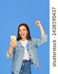 Excited gen z Latin girl looking at smartphone, happy Hispanic teen customer screaming celebrating win receiving good news using mobile cell phone winning online standing on blue background. Vertical.