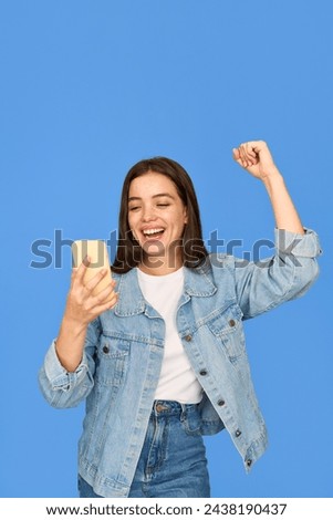Excited gen z Latin girl looking at smartphone, happy Hispanic teen customer screaming celebrating win receiving good news using mobile cell phone winning online standing on blue background. Vertical.