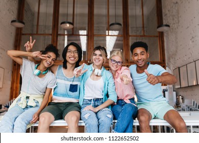 Excited girl in blue shirt showing peace sign enjoying friend's company in good day. Indoor portrait of glad international students fooling around for photo and laughing. - Powered by Shutterstock