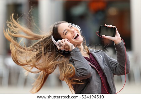 Excited girl dancing and listening music with headphones and smart phone in the street with hair moving